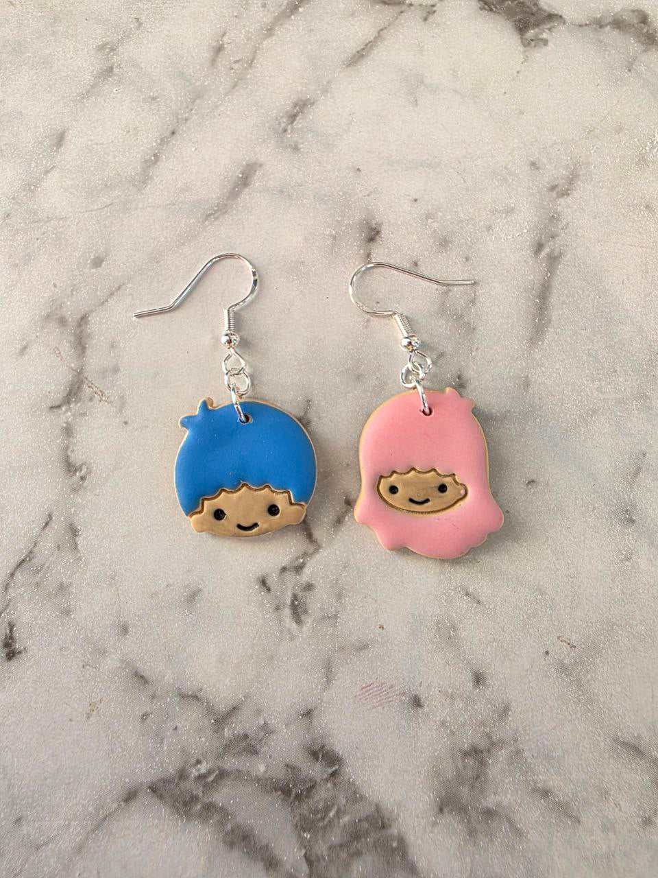 Lala and Kiki mismatched earrings – one with blue cartoon face and one with pink hooded character, handmade from polymer clay.