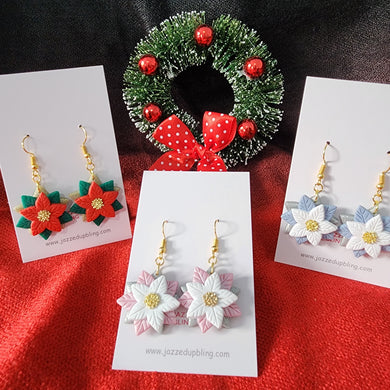 Handmade poinsettia Christmas earrings in red, pink, and blue with gold-colored hooks, displayed with festive wreath background.